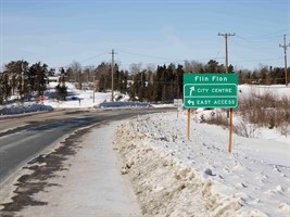 An image of a highway with a sign on the right side, indicating directions for Flin Flon City Centre and East access.
